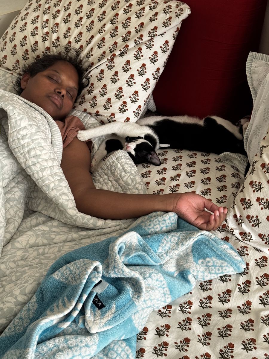 Prema asleep in bed with a black and white cat pressed against her shoulder and another stretched out beside her