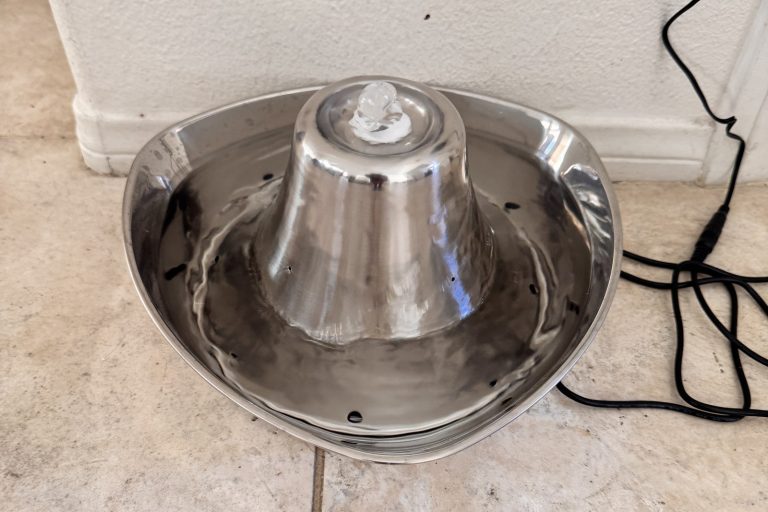 An open stainless steel cat fountain beside a wall on a tile floor